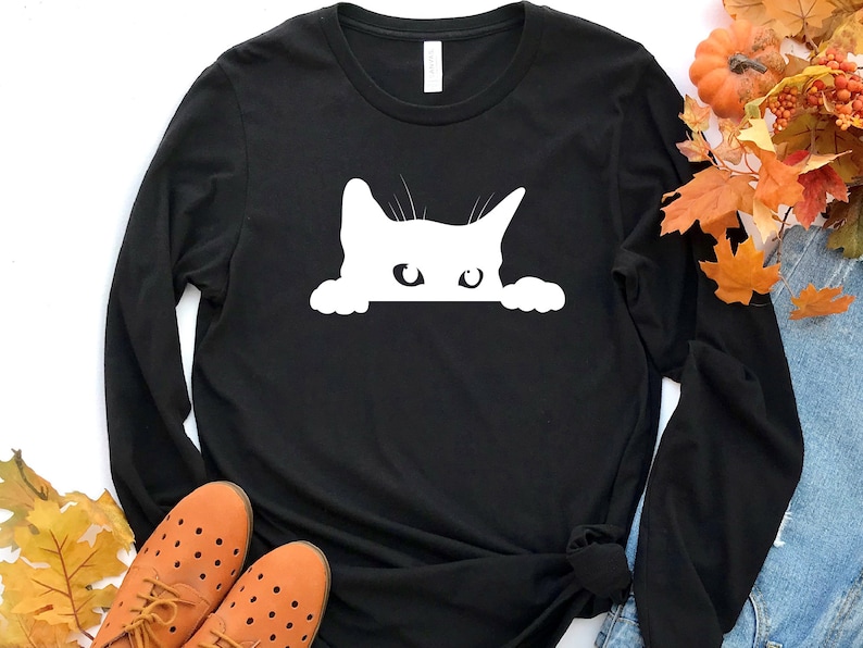 May include: Black long-sleeved t-shirt with a white silhouette of a cat peeking out from behind the fabric.