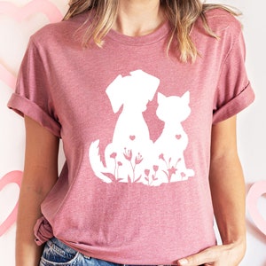 May include: A pink t-shirt with a white silhouette of a dog and cat sitting together. The design includes small white flowers and hearts.