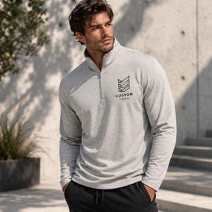 May include: A light gray long-sleeve pullover with a quarter-zip neckline. The shirt features a black logo and the text "CUSTOM LOGO" on the chest. The model is wearing black pants. The pullover is made of a soft, comfortable material.