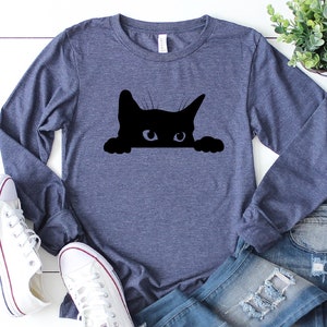 May include: A navy blue long-sleeved t-shirt featuring a black silhouette of a cat peeking over the top. The cat has white eyes.