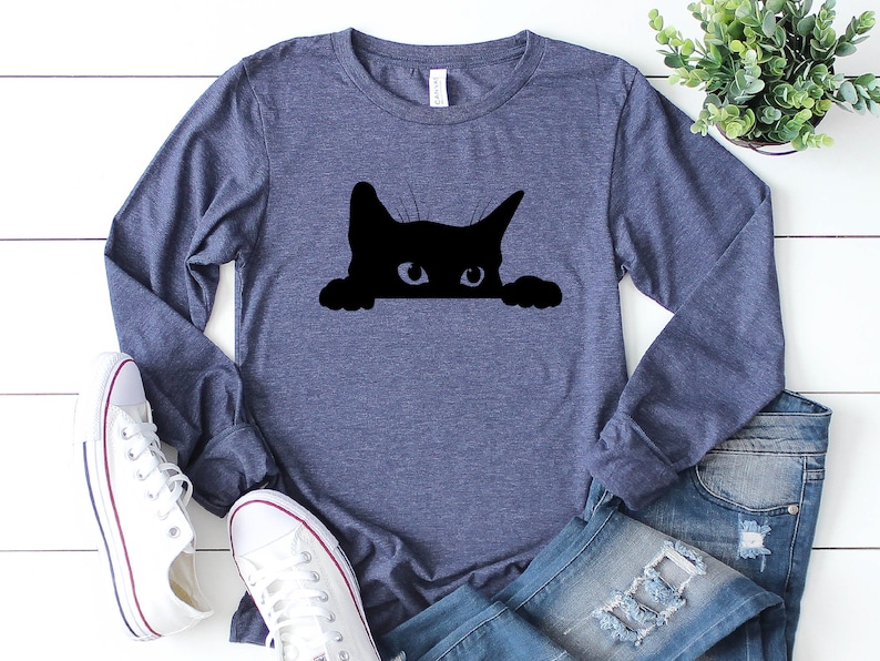 May include: A navy blue long-sleeved t-shirt featuring a black silhouette of a cat peeking over the top. The cat has white eyes.