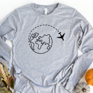 May include: A gray long-sleeved shirt with a black graphic of a plane flying around the world. The graphic is a simple line drawing of a globe with a plane flying above it.