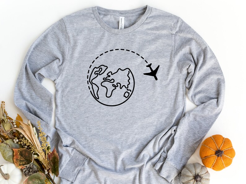May include: A gray long-sleeved shirt with a black graphic of a plane flying around the world. The graphic is a simple line drawing of a globe with a plane flying above it.