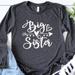 May include: A dark gray long-sleeved shirt with the text "Big Sister" in white lettering. The text is in a cursive font and includes a heart and arrow graphic.