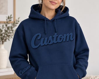May include: A navy blue hooded sweatshirt featuring the word Custom in a raised, tonal script font across the chest. The same word Custom is also embroidered in a smaller size on the right sleeve cuff. The hoodie includes a front kangaroo pocket.