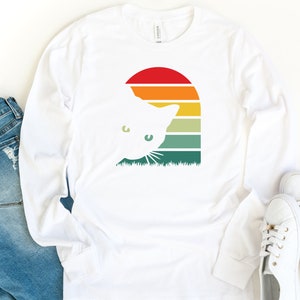 May include: White long-sleeved t-shirt with a retro sunset graphic featuring a cat peeking out from behind the sun. The graphic is in shades of orange, yellow, green, and blue.