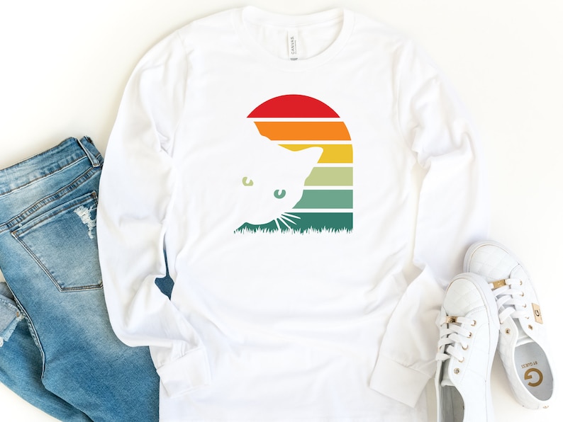 May include: White long-sleeved t-shirt with a retro sunset graphic featuring a cat peeking out from behind the sun. The graphic is in shades of orange, yellow, green, and blue.