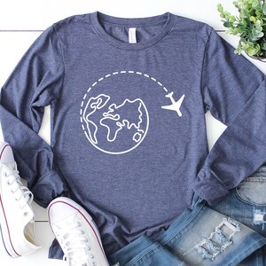 May include: A navy blue long-sleeved shirt with a white graphic of a globe and airplane. The shirt is laid out on a white wooden surface with a pair of white sneakers and blue jeans.