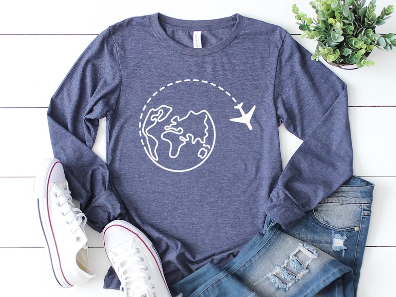 May include: A navy blue long-sleeved shirt with a white graphic of a globe and airplane. The shirt is laid out on a white wooden surface with a pair of white sneakers and blue jeans.