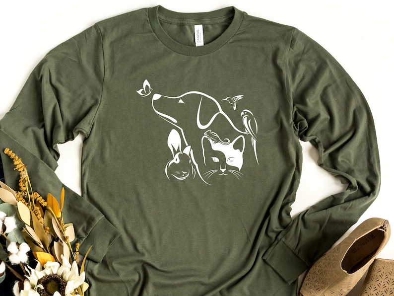Op de afbeelding: Een groen longsleeve t-shirt met een wit grafisch ontwerp van een hond, kat, konijn, vlinder en kolibrie.
