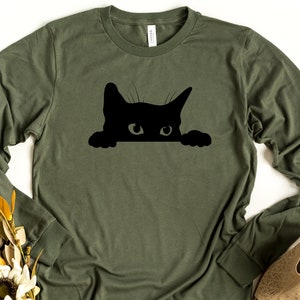 May include: A green long-sleeved t-shirt with a black silhouette of a cat peeking over the top.