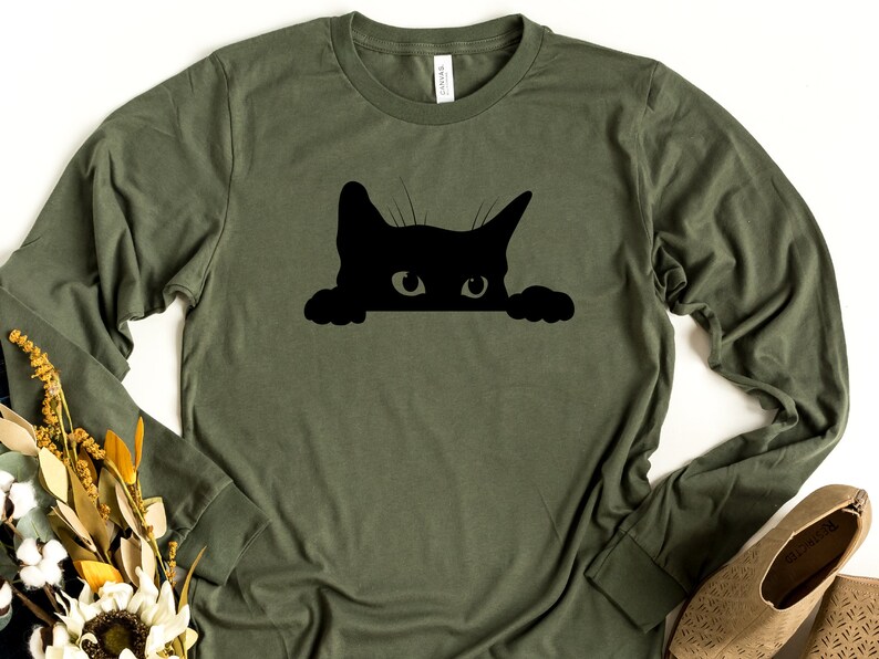 May include: A green long-sleeved t-shirt with a black silhouette of a cat peeking over the top.