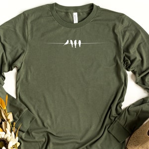 May include: A long-sleeved, olive green t-shirt with a white graphic of four birds perched on a wire.