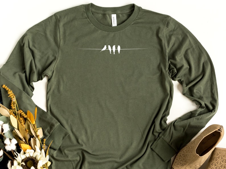 May include: A long-sleeved, olive green t-shirt with a white graphic of four birds perched on a wire.