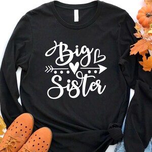 May include: Black long-sleeve shirt with white text that reads "Big Sister" with an arrow and hearts.