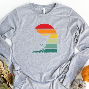 May include: A gray long-sleeved t-shirt with a retro-style graphic of a cat's head in front of a sunset. The graphic features a silhouette of a cat's head with green eyes, a red sun, and orange, yellow, green, and blue stripes.