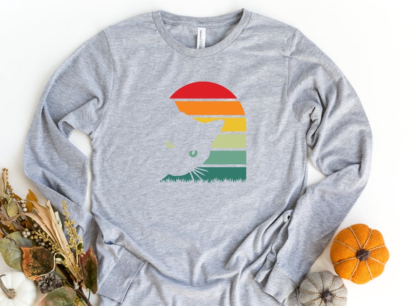 May include: A gray long-sleeved t-shirt with a retro-style graphic of a cat's head in front of a sunset. The graphic features a silhouette of a cat's head with green eyes, a red sun, and orange, yellow, green, and blue stripes.