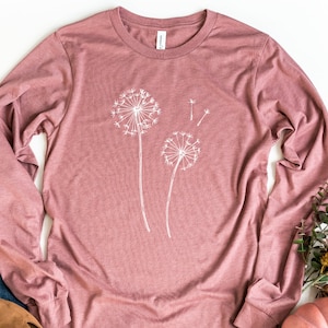 May include: A long-sleeved t-shirt in a dusty rose colour with a white graphic of two dandelions. The dandelions are stylised and have a delicate, whimsical look.