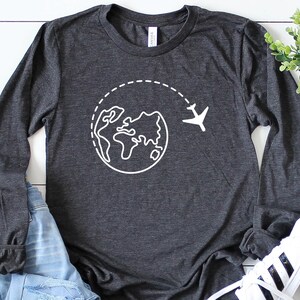 May include: A dark gray long-sleeved t-shirt with a white graphic of a globe with a plane flying around it. The shirt is laid out on a white wooden surface.