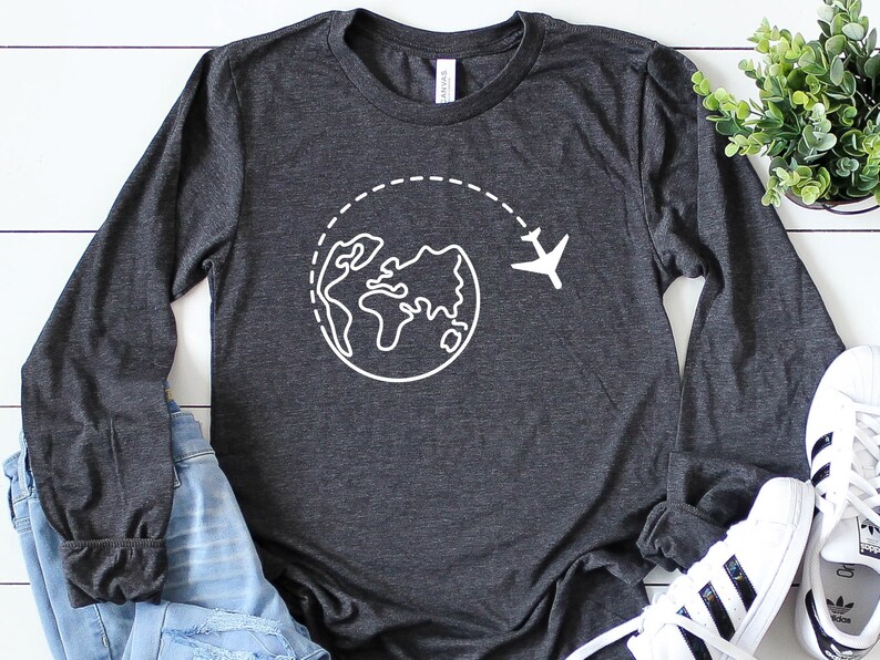 May include: A dark gray long-sleeved t-shirt with a white graphic of a globe with a plane flying around it. The shirt is laid out on a white wooden surface.