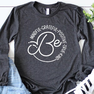 May include: A dark grey long-sleeved shirt with a white circle design that says "Be" in a cursive font. The circle is surrounded by the words "Mindful, Grateful, Positive, True, Kind".