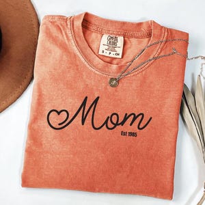 a red t-shirt with the word "Mom" printed on it, along with the year "Est 1965" below it. The t-shirt is folded and placed on a white surface, accompanied by a brown hat and some dried leaves.