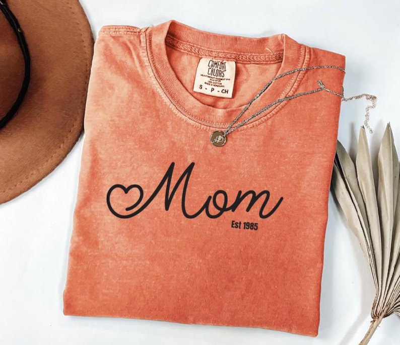 a red t-shirt with the word "Mom" printed on it, along with the year "Est 1965" below it. The t-shirt is folded and placed on a white surface, accompanied by a brown hat and some dried leaves.