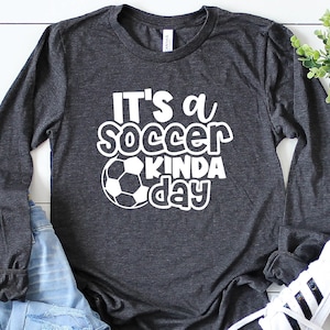 May include: Dark gray long-sleeve shirt with the text "It's a soccer kinda day" in white, along with a soccer ball graphic. The shirt is paired with blue jeans and white sneakers with black stripes.