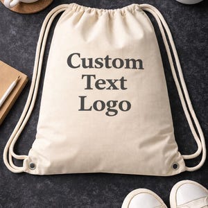May include: A beige drawstring backpack with the words "Custom Text Logo" printed in black. The bag has white drawstrings and metal eyelets. A pair of white sneakers is at the bottom.