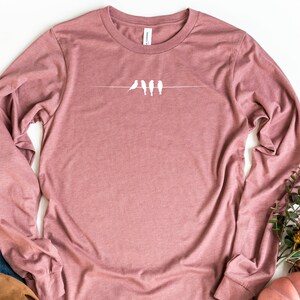 May include: A long-sleeved t-shirt in a dusty rose color with a white graphic of five birds perched on a wire.