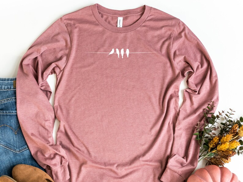 May include: A long-sleeved t-shirt in a dusty rose color with a white graphic of five birds perched on a wire.