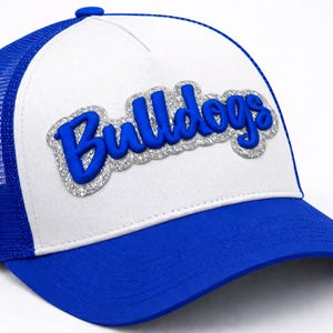 May include: A white and blue baseball cap with the word "Bulldogs" in blue, outlined with silver glitter. The cap has a blue brim and a blue mesh back.
