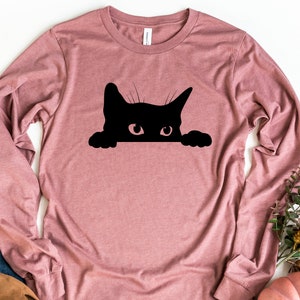 May include: A long-sleeved, mauve t-shirt featuring a black silhouette of a cat peeking over the top. The cat has a single, large eye visible.