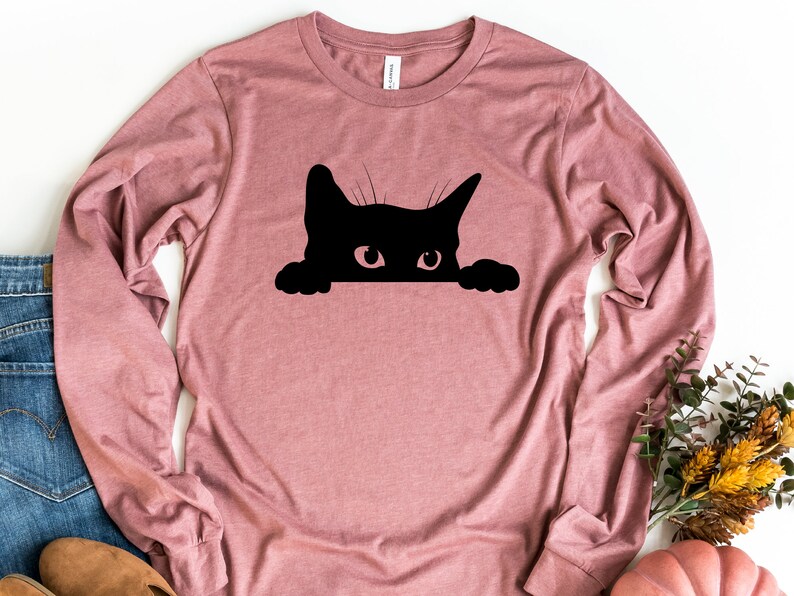 May include: A long-sleeved, mauve t-shirt featuring a black silhouette of a cat peeking over the top. The cat has a single, large eye visible.