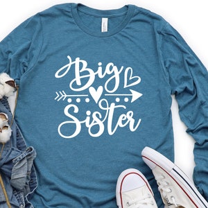 May include: A teal blue long-sleeved shirt with the text "Big Sister" in white lettering. The text is in a cursive font and includes a small heart and arrow graphic.