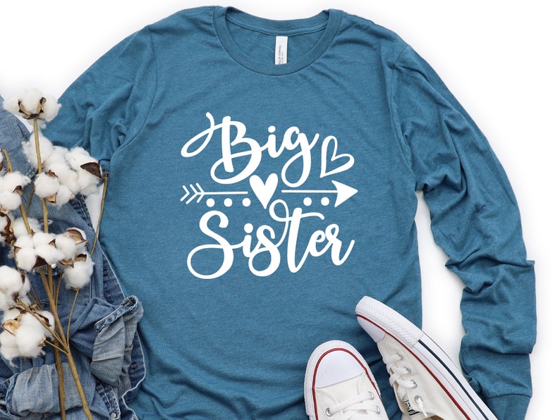 May include: A teal blue long-sleeved shirt with the text "Big Sister" in white lettering. The text is in a cursive font and includes a small heart and arrow graphic.