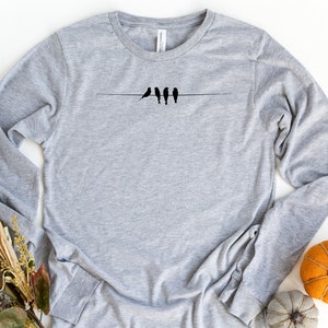 May include: A gray long-sleeved t-shirt with a simple design of four black birds perched on a wire.