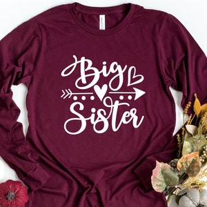 May include: A maroon long-sleeved shirt with the text "Big Sister" in white lettering. The text is stylized with a heart and arrow graphic.