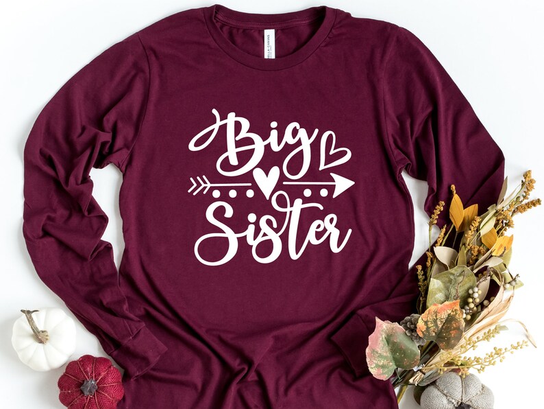 May include: A maroon long-sleeved shirt with the text "Big Sister" in white lettering. The text is stylized with a heart and arrow graphic.