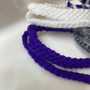 May include: Two strands of purple yarn and one strand of white yarn are coiled together. The yarn is soft and fluffy.