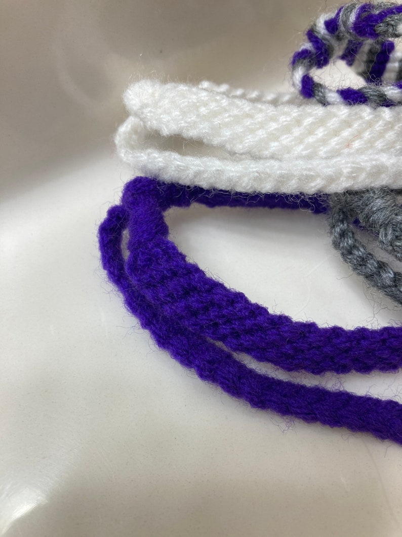 May include: Two strands of purple yarn and one strand of white yarn are coiled together. The yarn is soft and fluffy.