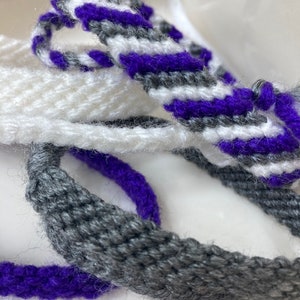 May include: Three woven bracelets in purple, gray, and white. The bracelets are made with a simple weave pattern and have a fuzzy texture.