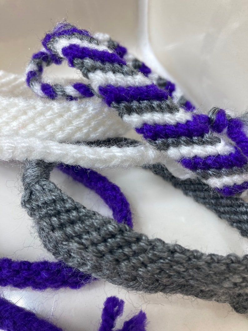 May include: Three woven bracelets in purple, gray, and white. The bracelets are made with a simple weave pattern and have a fuzzy texture.