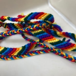 May include: A close-up of a colorful friendship bracelet made with woven threads in shades of red, orange, yellow, green, blue, and purple.