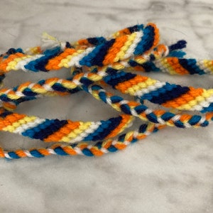 May include: A multi-coloured friendship bracelet with blue, orange, white and yellow threads woven together in a diagonal pattern.