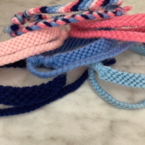 Pode incluir: Uma coleção de pulseiras de amizade coloridas em tons de rosa, azul e azul marinho. As pulseiras são feitas com fio tecido e estão amarradas juntas em um nó.