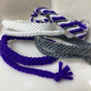 May include: A set of four woven friendship bracelets in purple, white, and gray. The bracelets are made with a simple, but intricate, knotting technique.