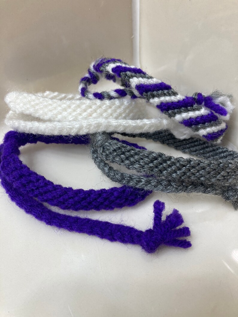 May include: A set of four woven friendship bracelets in purple, white, and gray. The bracelets are made with a simple, but intricate, knotting technique.