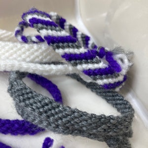 May include: Three friendship bracelets in white, gray, and purple. The bracelets are woven with a simple pattern and have frayed ends.