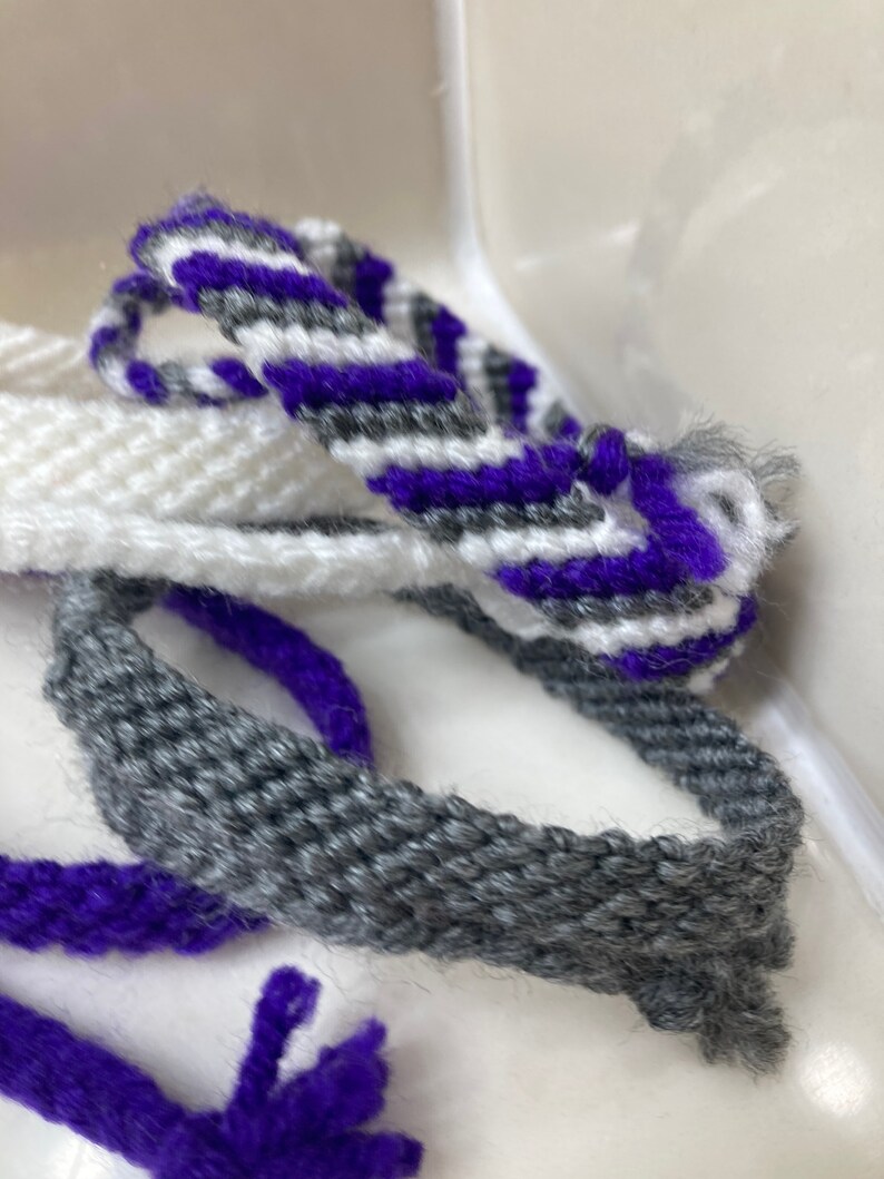 May include: Three friendship bracelets in white, gray, and purple. The bracelets are woven with a simple pattern and have frayed ends.
